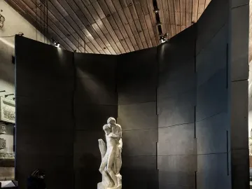 Michelangelo’s Rondanini Pietà displayed on a pedestal in a dim museum gallery, with a visitor standing nearby.