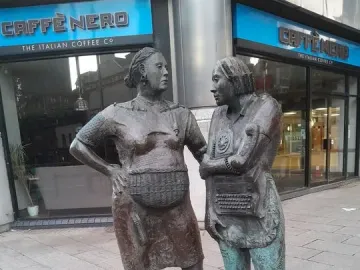 Monument to the Unknown Woman Worker with two bronze women standing on a paved street outside a Caffè Nero.