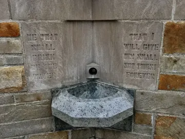 Stone holy water font at the National Shrine of the Little Flower with a metal spigot and carved inscription.