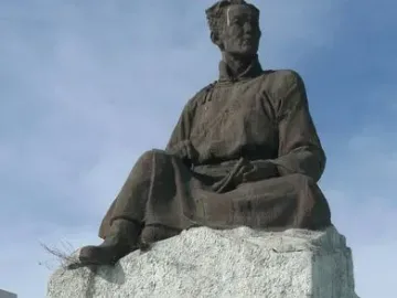 Natsagdorj Monument showing a weathered bronze-colored seated figure on a stone pedestal with engraved inscriptions and a floral wreath at the base.