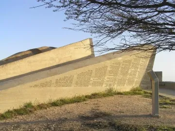 The Negev Brigade Monument, a large angular concrete structure covered with inscriptions, stands on rocky ground beneath a clear blue sky.