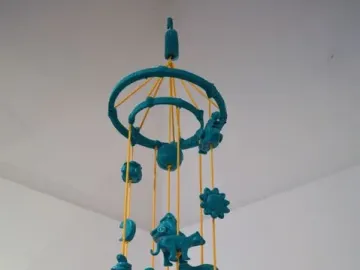 Teal cartonería mobile with small figures hanging from yellow strings, including animals, celestial shapes, and abstract forms.