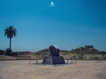 Lion of Babylon sculpture on a modern pedestal in a paved area with palm trees and distant hills behind it.