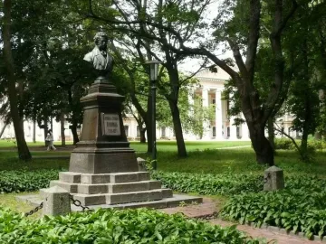 Bust of Pushkin on a tall stone pedestal in a park, with trees and a columned building in the background.