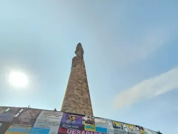 The Roundabout of the Disappeared monument rising against a bright blue sky, with posters and a disappeared-person flyer covering its base.