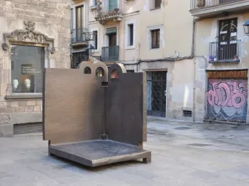 Topos V, a large dark L-shaped sculpture with arched cut-outs, standing on a stone-paved plaza in Barcelona.