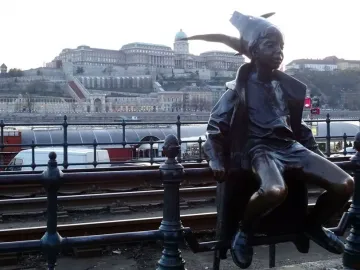 Little Princess bronze statue of a child in a coat and pointed cap sitting on a railing with a river and hillside buildings behind it.