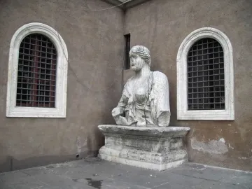 Madama Lucrezia stone bust on an ornate pedestal beside arched windows in a weathered wall.