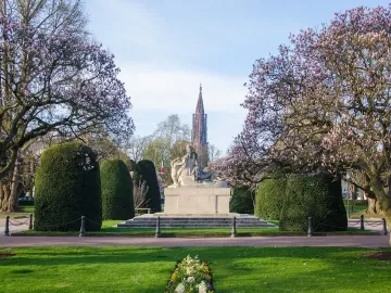 Stone war memorial sculpture with several figures on a stepped base, framed by pink-blossomed trees and trimmed hedges.