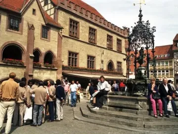 Gänseliesel Fountain in a town square, with a statue beneath an ornate dark spire and people sitting on the steps.