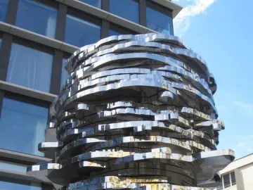 The Head of Franz Kafka sculpture with stacked, mirrored metal panels reflecting nearby buildings and blue sky.