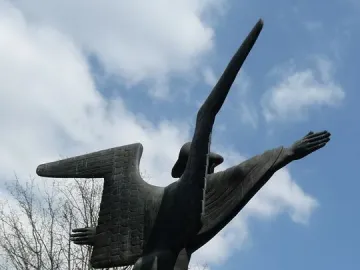 The Angel of Peace sculpture shown from below against a blue sky with clouds, with one arm extended and a pointed form rising beside it.
