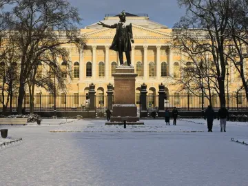 Pushkin statue on a pedestal in a snowy park, with a yellow building and bare winter trees in the background.