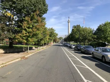 Trenton Battle Monument rises in the distance beside an American flag along a tree-lined street.