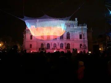 1.26 illuminated mesh sculpture glowing in bands of orange, yellow, and blue above a crowd near the Rudolfinum at night.