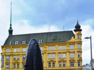 The Brno astronomical clock stands in a paved square with a yellow building and spire behind it.