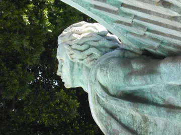 Close-up of the Monumento a Franco showing a verdigris-covered statue with wavy hair, draped clothing, and textured wings.