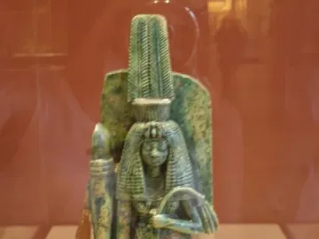 Standing statue of Queen Tiye with a feathered headdress, long wig, and patterned gown, holding a staff-like object.