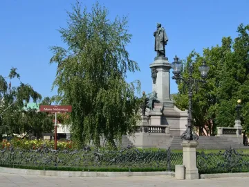 Adam Mickiewicz Monument with a bronze cloaked statue on a stone pedestal in Warsaw, framed by tree leaves and rose bushes.