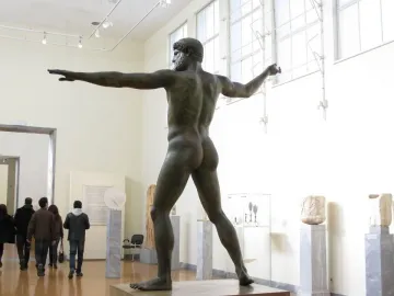Artemision Bronze shown from the back and left side in a museum gallery, with one arm extended and the other raised.