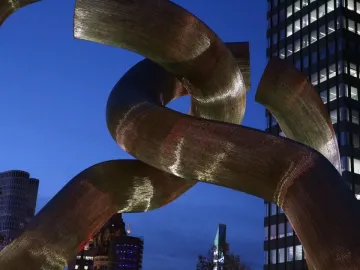 A golden sculpture of intertwined metallic tubes stands against a twilight sky with city buildings in the background.