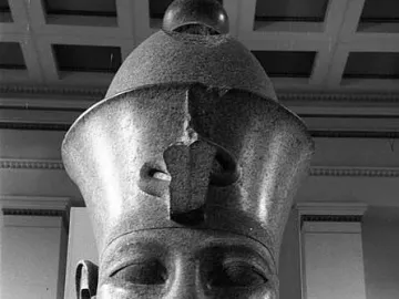 Colossal head of Amenhotep III with a tall crown and damaged neck, displayed on a base.