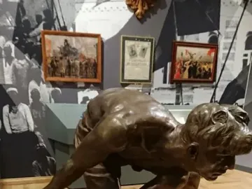 Bronze sculpture of a muscular man bent over gripping a large stone, displayed on a pedestal in a museum.