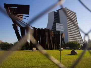 Monument to the Azoreans seen behind a chain-link fence, with a modern building and a restoration sign in the background.