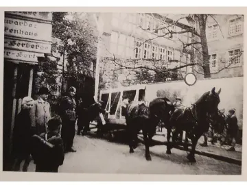 Horse-drawn covered vehicle moving through a street with several people and buildings in the background.