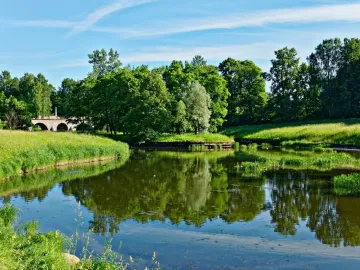 A slender monument to Elena Pavlovna stands near a multi-arched stone bridge over calm water surrounded by green trees.