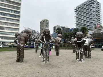 A-maze-ing Laughter statues standing on sandy ground with city buildings and trees behind them.