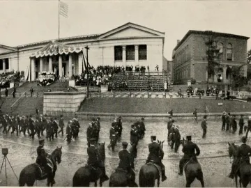 A large parade of uniformed men marches along a wet street past a decorated building, with the Charles Devens statue covered by an American flag and crowds gathered nearby.