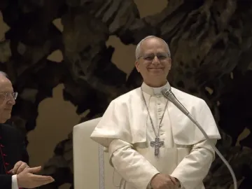 An elderly cleric in white vestments with glasses and a crucifix necklace smiles beside a microphone, with another cleric partly visible at left.