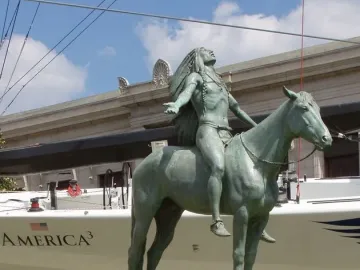 Appeal to the Great Spirit bronze statue of a Native American rider on horseback with arms outstretched, set against a sailboat and stone building.