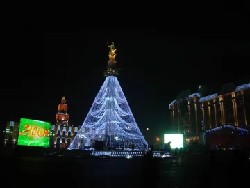 The Freedom Monument topped with a gilded figure, surrounded by bright blue lights forming a towering cone at night.