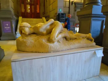 Malgré Tout, a reclining unclothed female sculpture with bound ankles on a textured base.