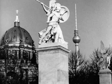 Athena Protects the Young Hero statue on a pedestal with pedestrians nearby, with the Berlin Cathedral and TV tower in the background.