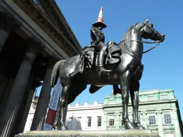 Duke of Wellington bronze equestrian statue with a traffic cone on the rider’s hat, set against a grand building under a blue sky.