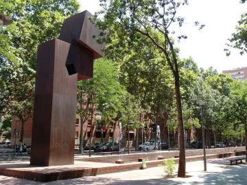 El Llarg Viatge, a tall dark abstract sculpture with a square opening beside a fountain and trees.