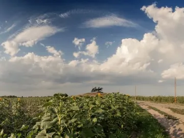 The Legendary Tachanka monument rises on a grassy mound beyond green fields and sunflowers under a dramatic blue sky.
