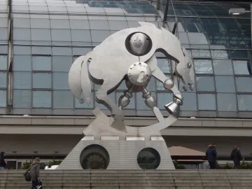 Rolling Horse sculpture beside a modern station with a vaulted glass roof, shown on a wedge-shaped pedestal with round porthole windows.