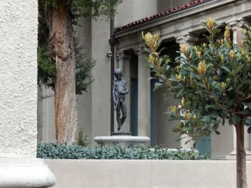Spirit of Spanish Music bronze statue of a boy holding a long pipe, framed by foliage and a courtyard building.