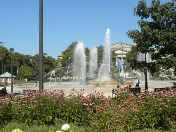 Swann Memorial Fountain rises behind a patch of pink flowers, with water shooting high from the circular basin.