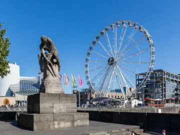 Tauzieher statue of a man securing a thick rope around a bollard, with a Ferris wheel and waterfront buildings in the background.