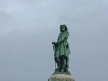 The Vercingetorix Monument, a bronze statue on a tall stone pedestal, stands on a grassy hill beneath an overcast sky.