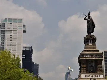 Cuauhtémoc Monument with a bronze figure in a feathered headdress on an ornate pedestal, set against modern buildings and trees.