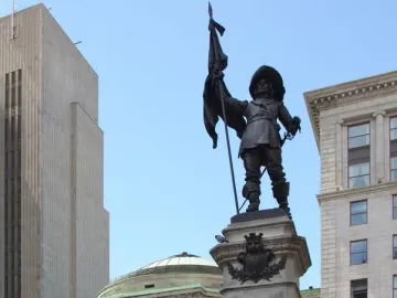 Maisonneuve Monument with a bronze figure holding a flag and sword atop a stone pedestal inscribed “MAISONNEUVE 1642,” with smaller figures below and city buildings in the background.
