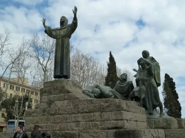 Monument to Saint Francis of Assisi with dark bronze figures on a stone plinth, including a hooded figure raising his hands toward the sky.