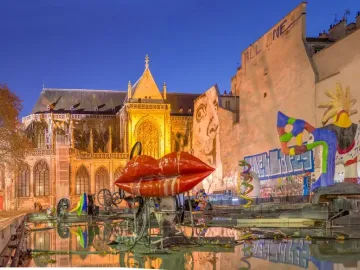 Stravinsky Fountain with colorful abstract sculptures, including red lips and a bird-like figure, beside an illuminated Gothic building at dusk.