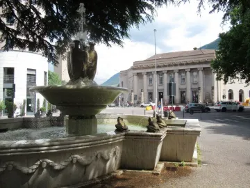 The Fountain of the Frogs stone fountain sprays water in a city square with a large classical building in the background.